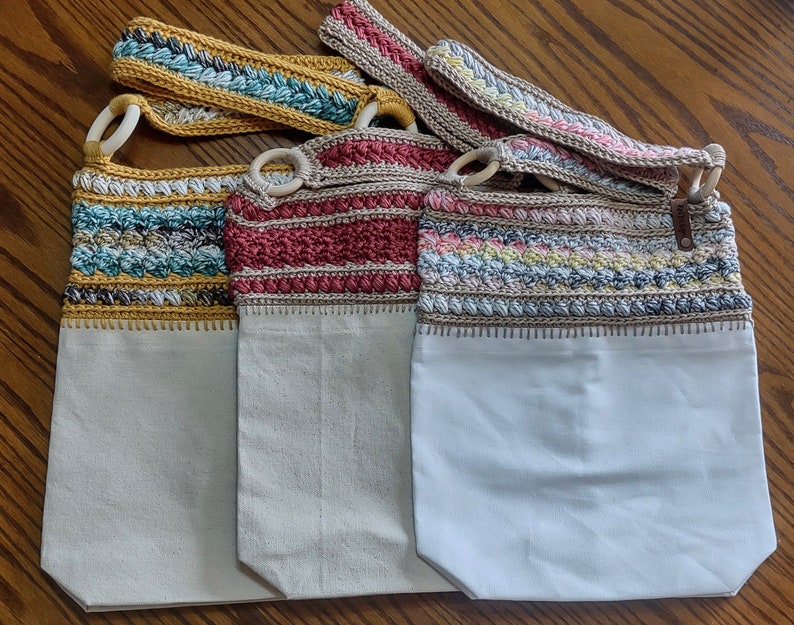 May include: Three crocheted tote bags with different coloured patterns. The bags are made with a white canvas base and have a crocheted strap.