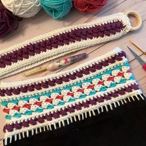 May include: A crocheted bag with a white, purple, teal and coral striped design. The bag has a white crocheted strap with a wooden ring.