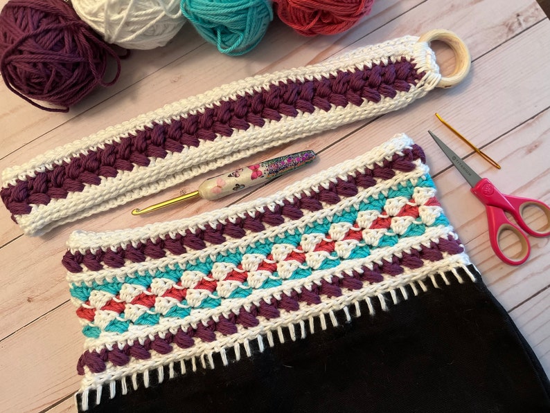 May include: A crocheted bag with a white, purple, teal and coral striped design. The bag has a white crocheted strap with a wooden ring.