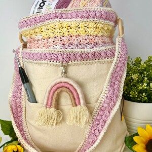 May include: A crocheted yarn tote bag with a pink and white rainbow keychain. The bag has a pink crocheted trim and a canvas bottom with a pocket. The bag is filled with yarn.