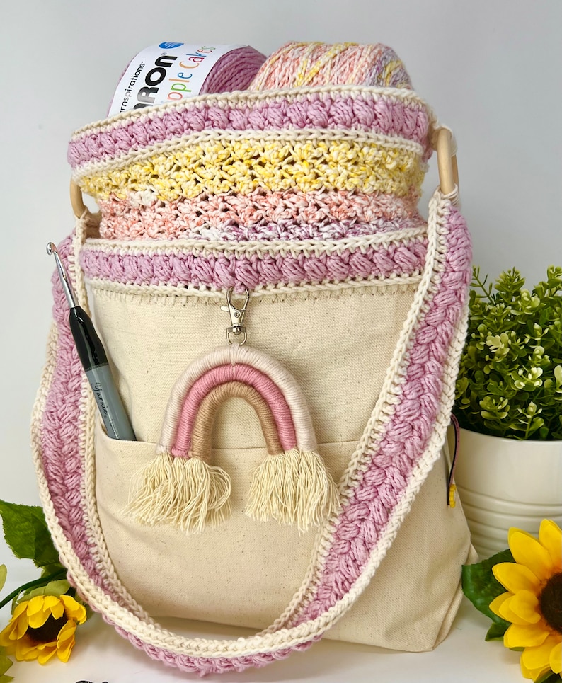 May include: A crocheted yarn tote bag with a pink and white rainbow keychain. The bag has a pink crocheted trim and a canvas bottom with a pocket. The bag is filled with yarn.