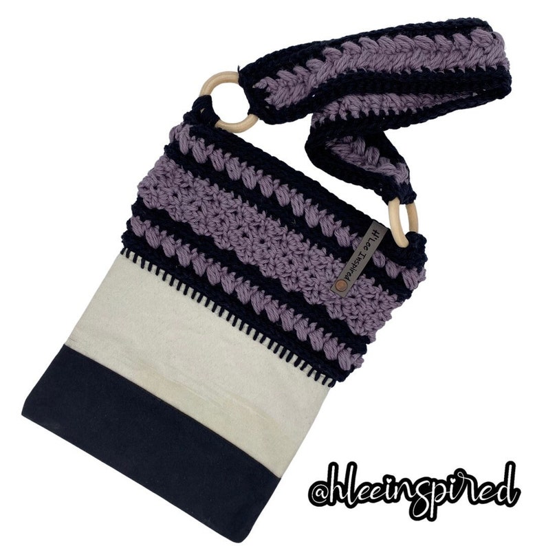 May include: A purple and black crocheted crossbody bag with a wooden ring handle. The bag has a white canvas lining and a black fabric base. The bag has a small brown tag with the text "Hilee Inspired" on it.