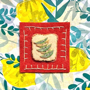May include: A felt art piece featuring a slice of toast with avocado slices on a red square, stitched with white thread. The background is a colorful pattern of lemons and leaves.