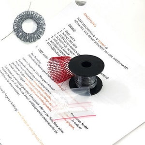 May include: A close-up of a craft kit for making jewelry. The kit includes a spool of black wire, a red mesh ring, a gray mesh ring, and instructions for making a donut-shaped necklace. The instructions are written in German.