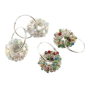 May include: Four pairs of silver hoop earrings with colorful crystal beads woven into the hoops. The earrings are arranged in a row on a white background.