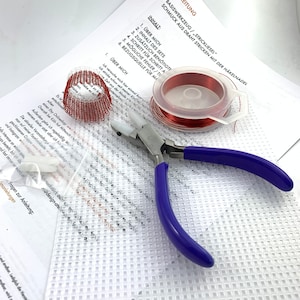 Könnte beinhalten: Nahaufnahme eines Bastelsets zum Schmuck herstellen. Das Set beinhaltet eine Spule roten Drahtes, eine Zange, ein Stück weißes Netzgewebe und eine Anleitung. Die Anleitung ist in deutscher Sprache verfasst und beinhaltet den Titel "Basiswerkzeug/Strickliesel" und "Schmuck aus Draht stricken mit der Häkelnadel".