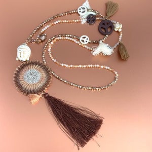 May include: A long, layered necklace with a brown and gold mandala pendant, a white Buddha charm, and peace sign charms. The necklace has a brown tassel and is made of beads and crystals.