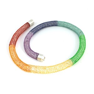 May include: A rainbow colored crystal necklace with silver end caps. The necklace is made of small crystals that are woven together to create a mesh-like effect. The colors of the necklace are orange, yellow, green, blue, and purple.