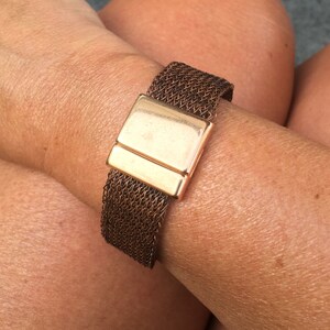 May include: A copper mesh bracelet with a gold clasp. The bracelet is on a person's wrist.