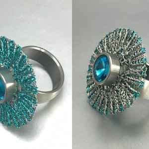 Puede incluir: Un anillo de plata con una piedra preciosa azul turquesa engastada en un centro de plata. El anillo está decorado con un diseño de malla de alambre de plata y azul turquesa.