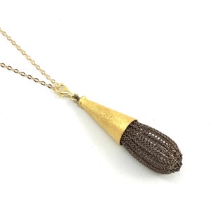 Puede incluir: Un collar dorado con un colgante. El colgante tiene un cono dorado texturizado y un elemento tejido en forma de lágrima, de color marrón oscuro. El collar tiene una delicada cadena y cierre dorados, sobre un fondo blanco.