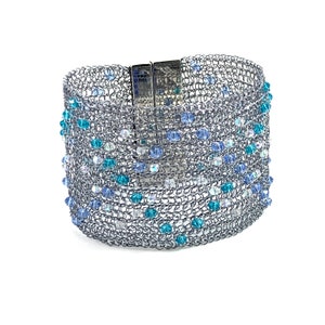 Pode incluir: Um bracelete de malha prateada com contas de cristal azul e transparente. O bracelete tem um fecho magnético.