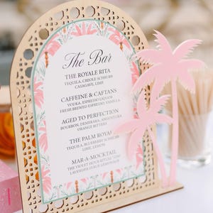 May include: An arched wooden menu sign with a laser-cut lattice border and a printed insert featuring pink palm trees. The text reads The Bar, The Royale Rita, Caffeine & Caftans, Aged to Perfection, The Palm Royal, and Mar-a-Mocktail. A pink palm tree cutout is attached to the right side.