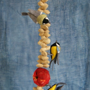 Op de afbeelding: Een vilten mobiel met drie vogels en een rode appel die hangen aan een touwtje met beige vilten cirkels. De vogels zijn geel, blauw en grijs met zwarte ogen. De mobiel hangt tegen een blauwe achtergrond.