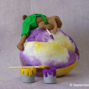 K&ouml;nnte beinhalten: Ein braunes und gr&uuml;nes Filztier, das eine Latzhose tr&auml;gt, malt ein lila, gelbes und wei&szlig;es Filzei mit einem Pinsel. Zwei kleine Farbdosen stehen auf dem Tisch.