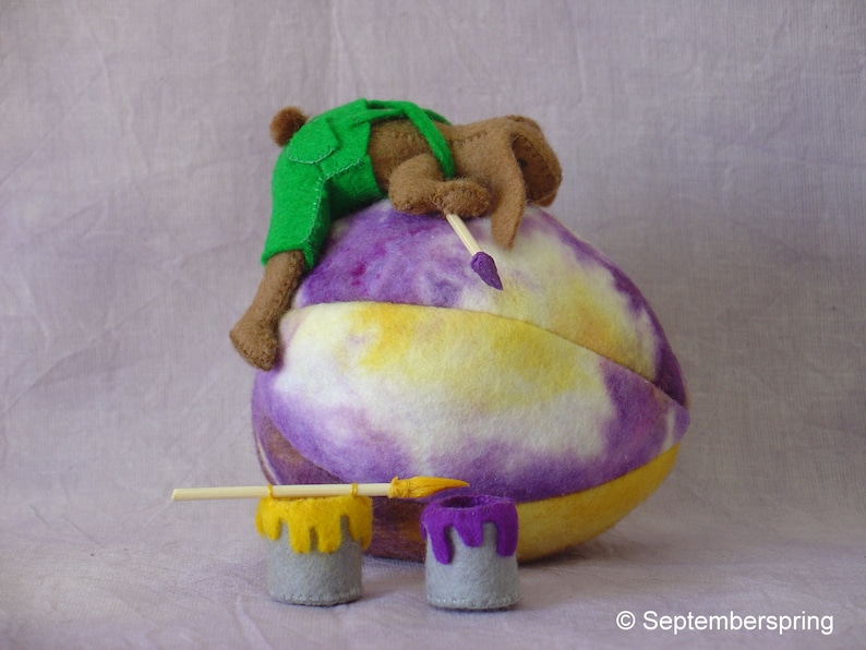 K&ouml;nnte beinhalten: Ein braunes und gr&uuml;nes Filztier, das eine Latzhose tr&auml;gt, malt ein lila, gelbes und wei&szlig;es Filzei mit einem Pinsel. Zwei kleine Farbdosen stehen auf dem Tisch.