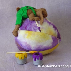 K&ouml;nnte beinhalten: Ein braunes und gr&uuml;nes Filztier, das eine Latzhose tr&auml;gt, malt ein gro&szlig;es lila und gelbes Ei mit einem Pinsel. Zwei kleine Farbt&ouml;pfe stehen auf dem Boden, einer gelb und einer lila.