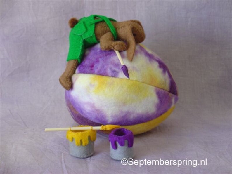 K&ouml;nnte beinhalten: Ein braunes und gr&uuml;nes Filztier, das eine Latzhose tr&auml;gt, malt ein gro&szlig;es lila und gelbes Ei mit einem Pinsel. Zwei kleine Farbt&ouml;pfe stehen auf dem Boden, einer gelb und einer lila.