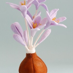 Könnte beinhalten: Ein Strauß aus hellvioletten Filzblumen mit gelben Zentren in einer braunen Filzvase. Die Vase hat die Form einer Zwiebel.