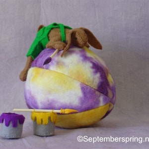 K&ouml;nnte beinhalten: Ein braunes und gr&uuml;nes Pl&uuml;schtier malt ein gro&szlig;es lila und gelbes Batik-Ei mit einem Pinsel. Zwei kleine Farbt&ouml;pfe mit lila und gelber Farbe befinden sich im Vordergrund.