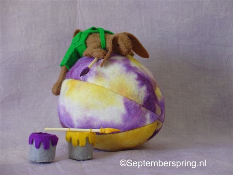 K&ouml;nnte beinhalten: Ein braunes und gr&uuml;nes Pl&uuml;schtier malt ein gro&szlig;es lila und gelbes Batik-Ei mit einem Pinsel. Zwei kleine Farbt&ouml;pfe mit lila und gelber Farbe befinden sich im Vordergrund.