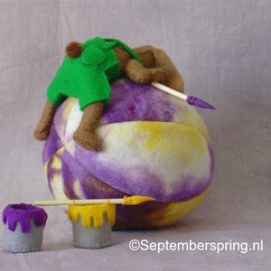 K&ouml;nnte beinhalten: Ein lila-gelber Filzkugel mit einem braunen und gr&uuml;nen Filzb&auml;r, der einen gr&uuml;nen Filzhut tr&auml;gt, der darauf sitzt. Zwei kleine lila-gelbe Farbdosen befinden sich im Vordergrund mit einem Holzpinsel.