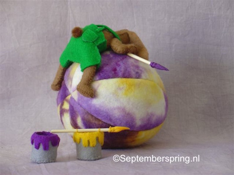 K&ouml;nnte beinhalten: Ein lila-gelber Filzkugel mit einem braunen und gr&uuml;nen Filzb&auml;r, der einen gr&uuml;nen Filzhut tr&auml;gt, der darauf sitzt. Zwei kleine lila-gelbe Farbdosen befinden sich im Vordergrund mit einem Holzpinsel.