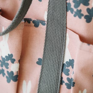 May include: Close-up of a pink canvas tote bag with a grey strap. The bag has a white rabbit and clover leaf pattern.