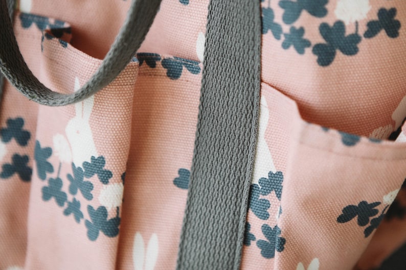 May include: Close-up of a pink canvas tote bag with a grey strap. The bag has a white rabbit and clover leaf pattern.