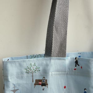 May include: A light blue tote bag with grey fabric handles. The bag has a white and light blue patterned design with illustrations of people, trees, and a fountain.