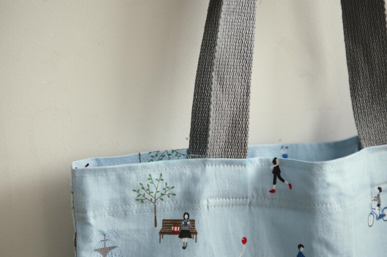 May include: A light blue tote bag with grey fabric handles. The bag has a white and light blue patterned design with illustrations of people, trees, and a fountain.