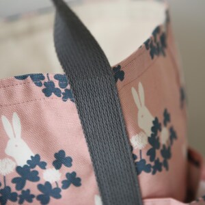 May include: A pink canvas tote bag with a black strap. The bag features a white rabbit and clover leaf design.