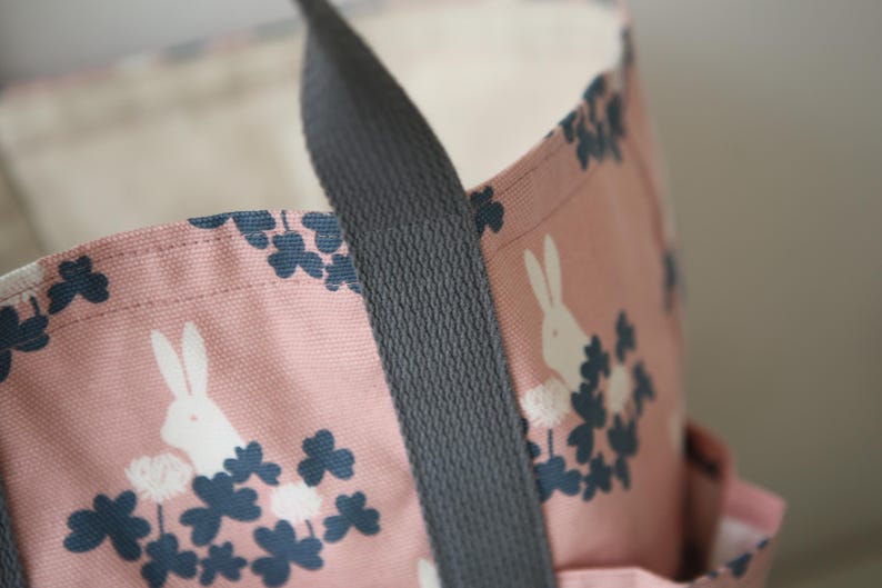 May include: A pink canvas tote bag with a black strap. The bag features a white rabbit and clover leaf design.