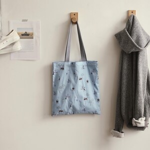 May include: A light blue tote bag with a white and black pattern hangs on a wooden hook. The bag has two straps and is made of a soft fabric. A grey hoodie hangs on a hook next to the bag.
