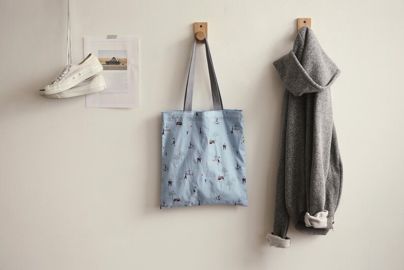 May include: A light blue tote bag with a white and black pattern hangs on a wooden hook. The bag has two straps and is made of a soft fabric. A grey hoodie hangs on a hook next to the bag.