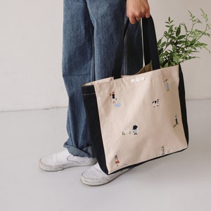May include: A large beige tote bag with black handles and a black trim. The bag has a white label with black text and is decorated with embroidered illustrations of people and animals.