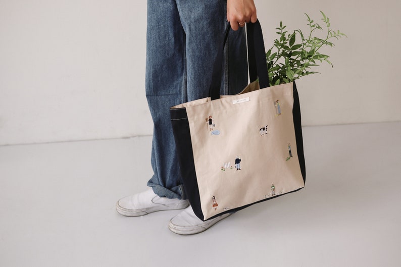 May include: A large beige tote bag with black handles and a black trim. The bag has a white label with black text and is decorated with embroidered illustrations of people and animals.