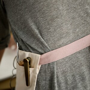 May include: A person wearing a grey shirt with a pink and white apron. The apron has a pocket with a brass clip holding a wooden tool.