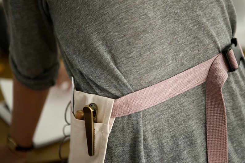 May include: A person wearing a grey shirt with a pink and white apron. The apron has a pocket with a brass clip holding a wooden tool.