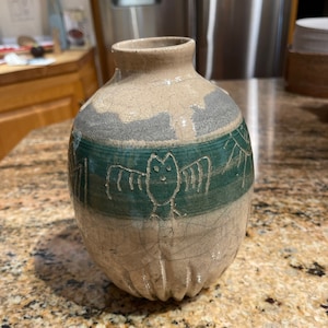 Puede incluir: Un jarrón de cerámica con un esmalte verde, gris y blanco. El jarrón tiene una superficie texturizada y un diseño de murciélago en la parte delantera.