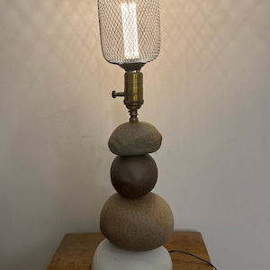 May include: A table lamp with a wire cage shade and a stacked stone base. The base is made of four round stones in different shades of brown and white.