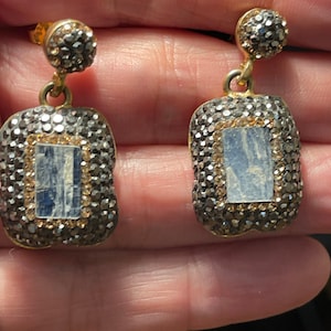 May include: A pair of silver and gold earrings with a rectangular blue stone set in the center. The earrings are surrounded by small clear and black stones.