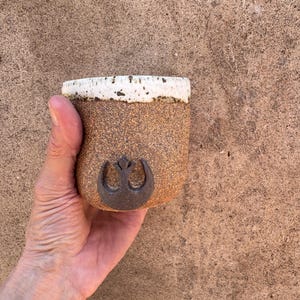 Könnte beinhalten: Eine handgefertigte, gesprenkelte braune Keramik-Tasse mit weißem Rand. Die Tasse zeigt ein dunkelbraunes Rebellenallianz-Symbol. Die Tasse wird in einer Hand vor einem braunen Hintergrund gehalten.