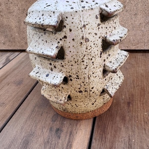 Puede incluir: Una maceta de cerámica moteada para fresas con múltiples niveles y aberturas. La maceta blanquecina tiene motas marrones y una superficie texturizada. La base es de color marrón rojizo. Diseñada para cultivar fresas u otras plantas pequeñas.