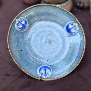 May include: A handmade ceramic plate with a blue and brown glaze. The plate features three raised blue emblems around the rim. The plate has a diameter of approximately 6 inches.