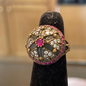 May include: A silver ring with a round, domed top. The top is decorated with a black and white geometric pattern and a single pink gemstone in the center. The ring is set on a black velvet ring holder.