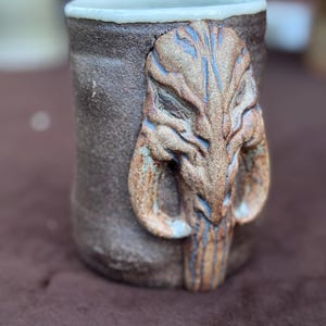 Könnte beinhalten: Handgefertigte Keramik-Tasse mit strukturierter brauner Außenseite und weißem Innenrand. Die Tasse zeigt ein detailliertes, geformtes Schädeldesign mit blauen und braunen Akzenten. Ein einzigartiges Trinkgefäß.