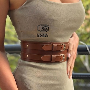 Puede incluir: Un cinturón de cuero marrón con dos correas y hebillas doradas. El cinturón se lleva sobre un vestido verde oliva claro. La imagen incluye el texto "LEATHER INSOMNIA".