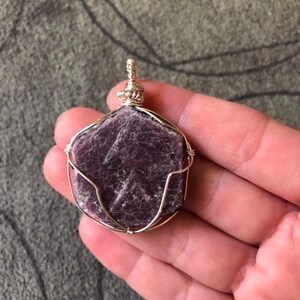 May include: A purple gemstone pendant wrapped in silver wire. The pendant is shaped like a hexagon and has a rough, natural texture.