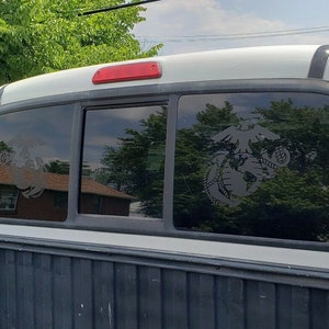 May include: A white pickup truck with a tinted rear window. The window has a black and white decal of the United States Marine Corps emblem.